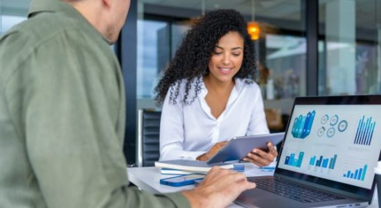 Business man and business woman in a meeting at the office. There is a laptop on the table  showing finance charts and graphs and the woman is holding a digital tablet. There is paperwork with finance chart and graphs.