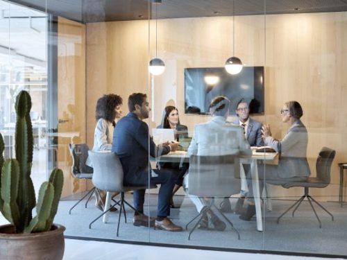 Senior businesswoman explaining strategy with colleagues in board room. Multi-ethnic professionals are discussing while sitting at desk in office. They are seen through glass.